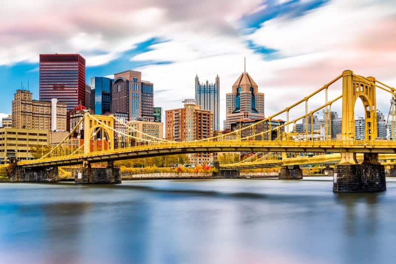 Yellow suspension bridge over a calm river with a city skyline in the background under a partly cloudy sky.