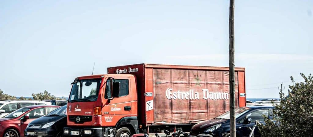 A red Estrella Damm truck is parked in a busy parking lot surrounded by various cars on a clear day.