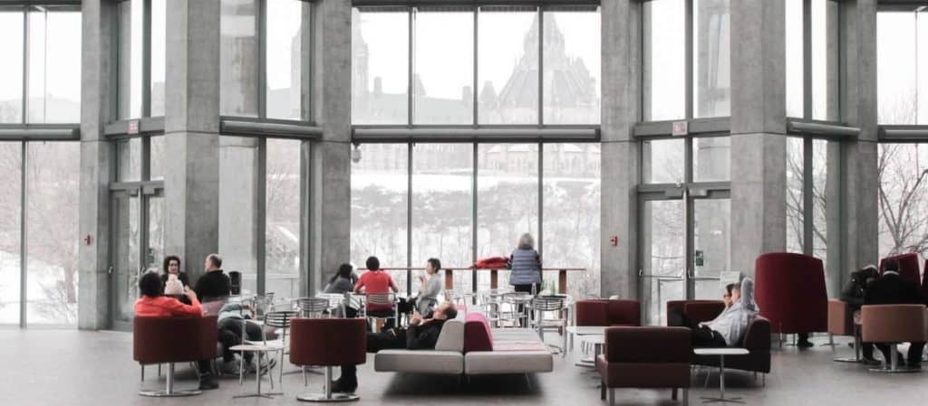 People sit and converse in a modern, glass-walled atrium with large windows offering a snowy outside view.