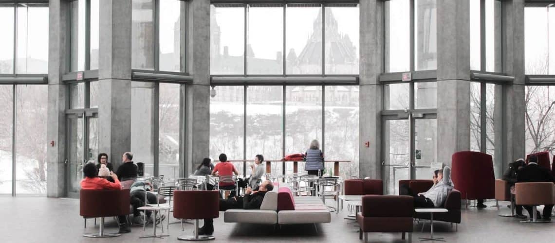 People sit and converse in a modern, glass-walled atrium with large windows offering a snowy outside view.