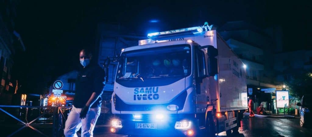A man wearing a mask walks past a parked SAMU Iveco emergency vehicle with flashing lights on a city street at night.