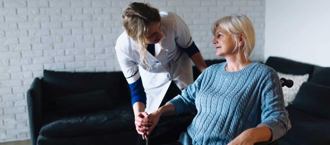 A caregiver in a white uniform leans over to assist an older woman sitting in a wheelchair in a living room.