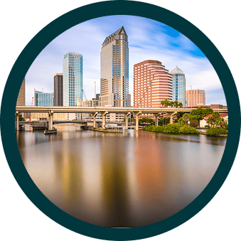 Circular image of a city skyline with tall buildings, a bridge, and a calm body of water in the foreground under a blue sky.