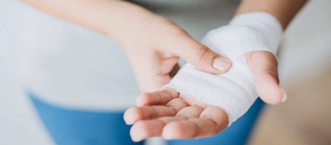 A person with a bandaged hand holds it gently, palm facing up. The bandage wraps around the palm and wrist.