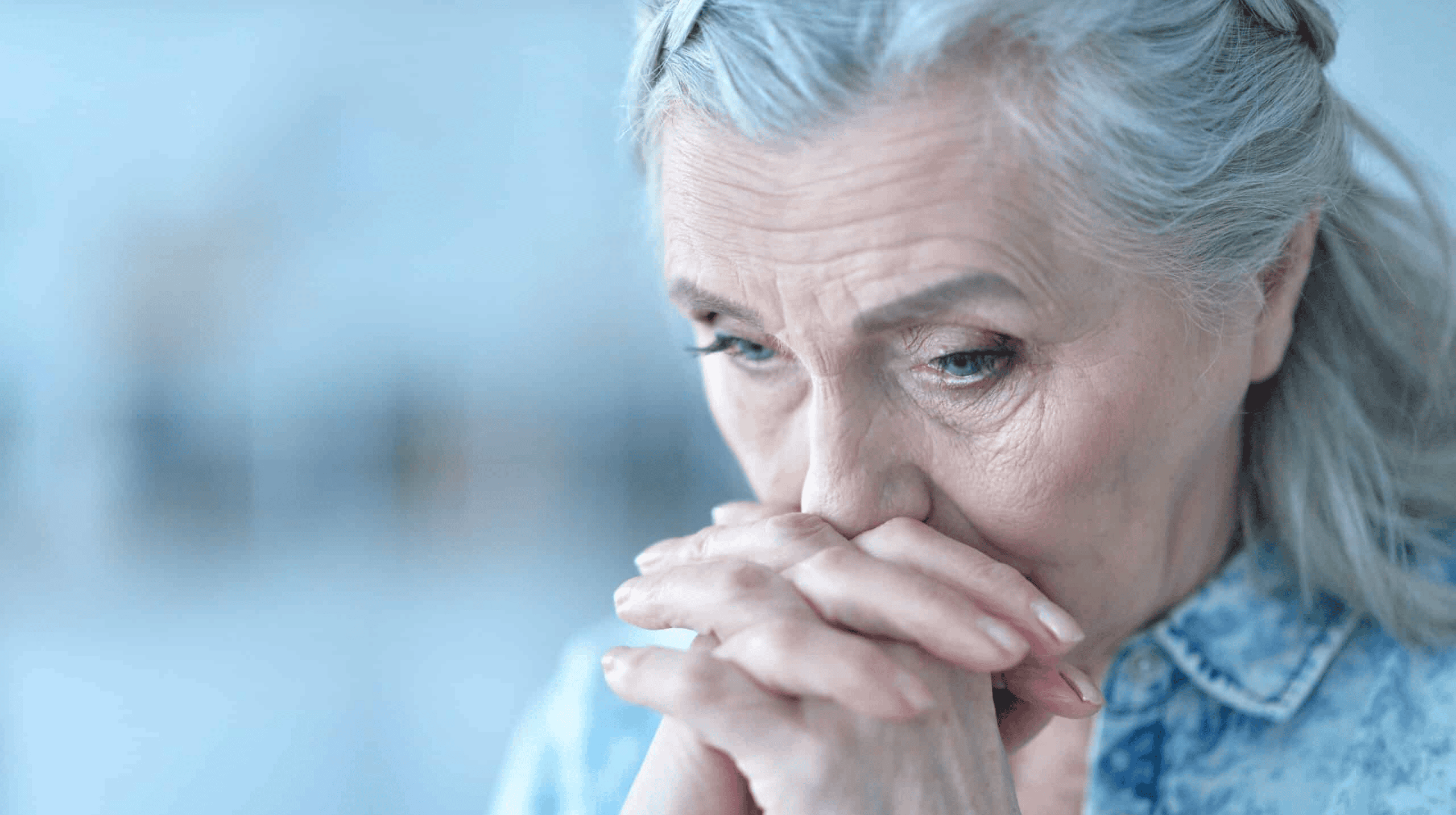 Elderly woman with gray hair, wearing a blue blouse, rests her chin on clasped hands, looking contemplative.