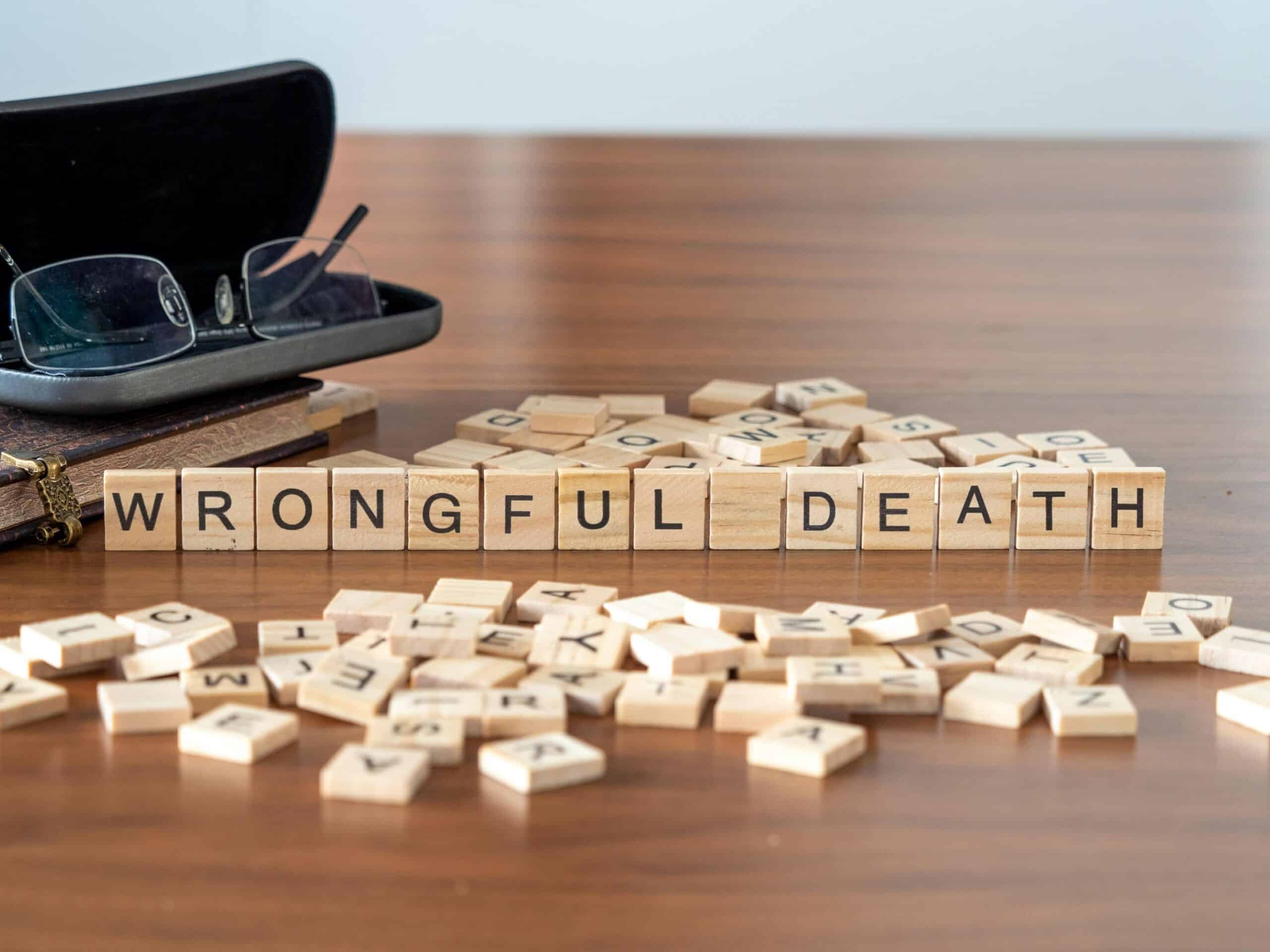 Wooden letter tiles spell "WRONGFUL DEATH" on a wooden table, surrounded by scattered tiles and a pair of glasses in a case.