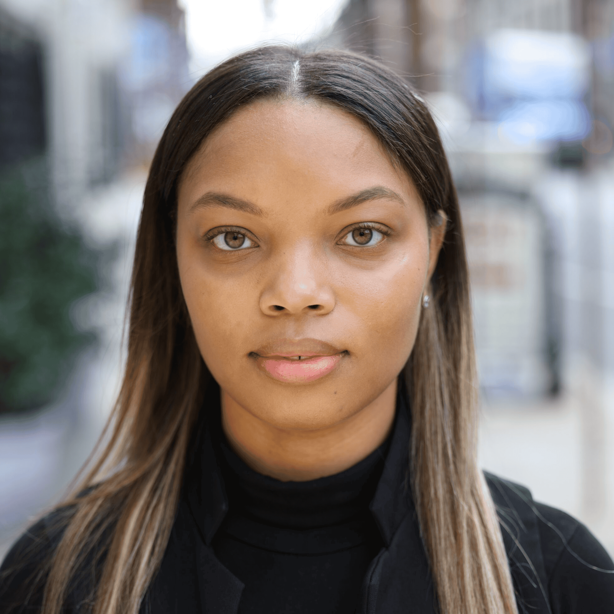 Woman with straight hair and a neutral expression standing outdoors, wearing a black top with a blurred urban background.