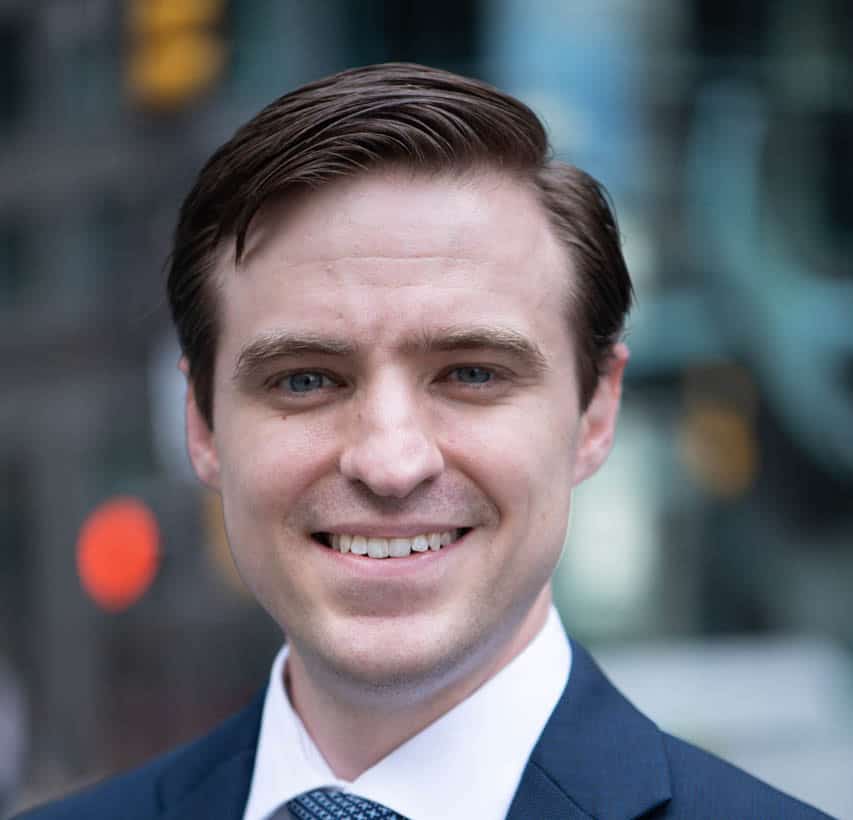 A man in a suit and tie smiling, with a blurred cityscape in the background.