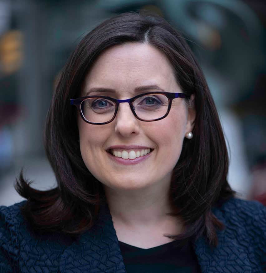 A woman with dark hair, wearing glasses and a black textured top, smiles at the camera. She is outdoors with a blurred background.