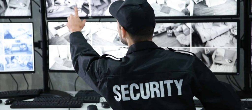 A security guard monitors multiple surveillance screens, pointing at one of the displays, in a control room.