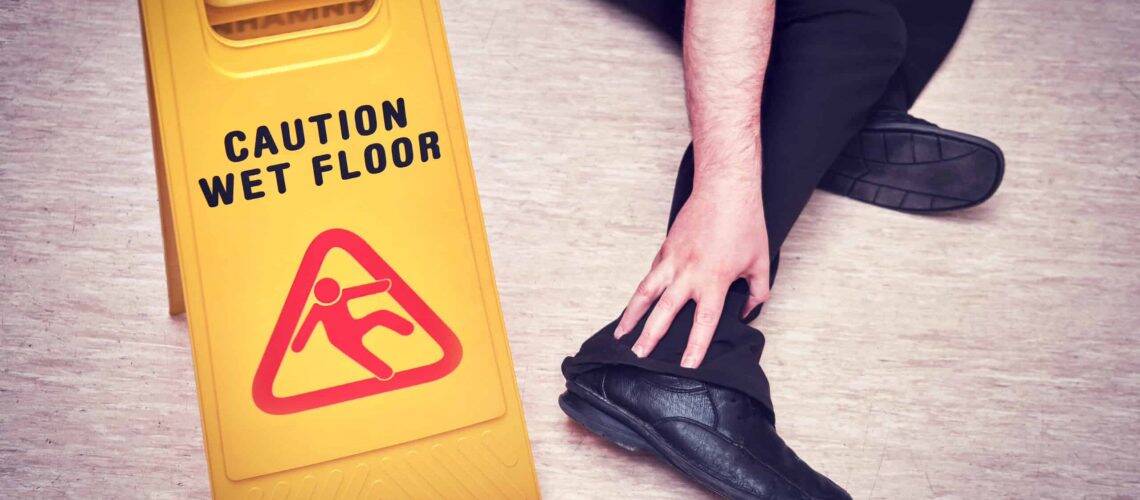 Person sitting on floor holding their leg near a yellow "Caution Wet Floor" sign.