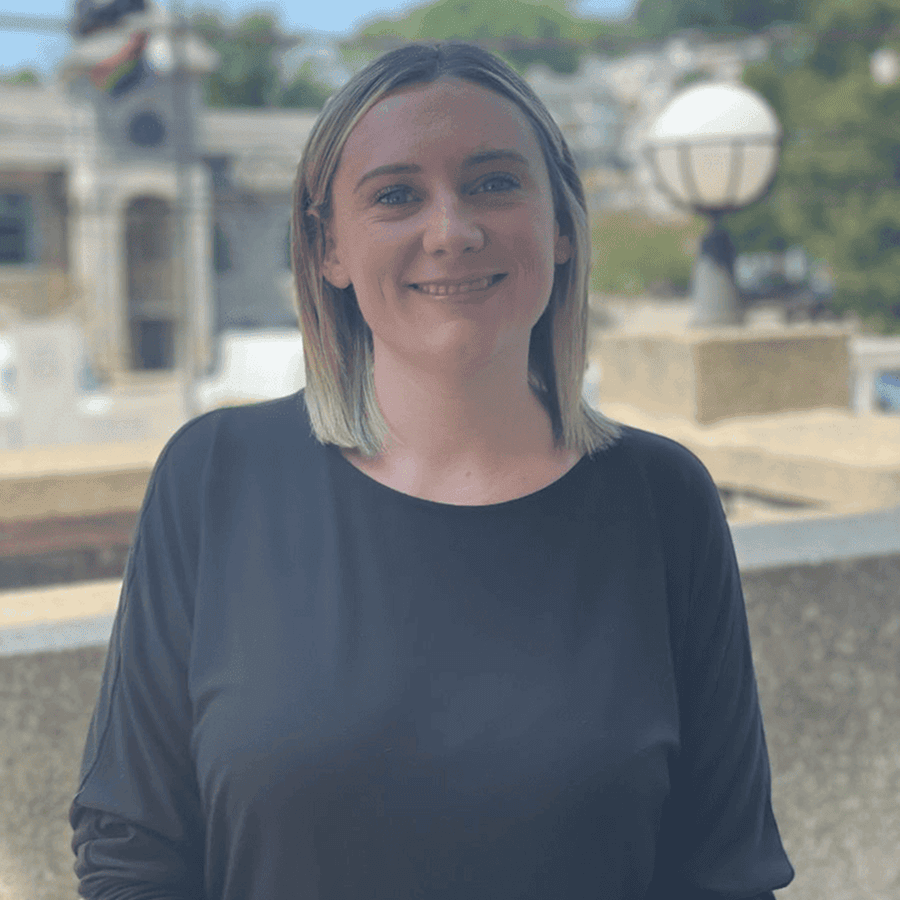 Person with shoulder-length hair stands outdoors, smiling, with a blurred background of buildings and greenery.