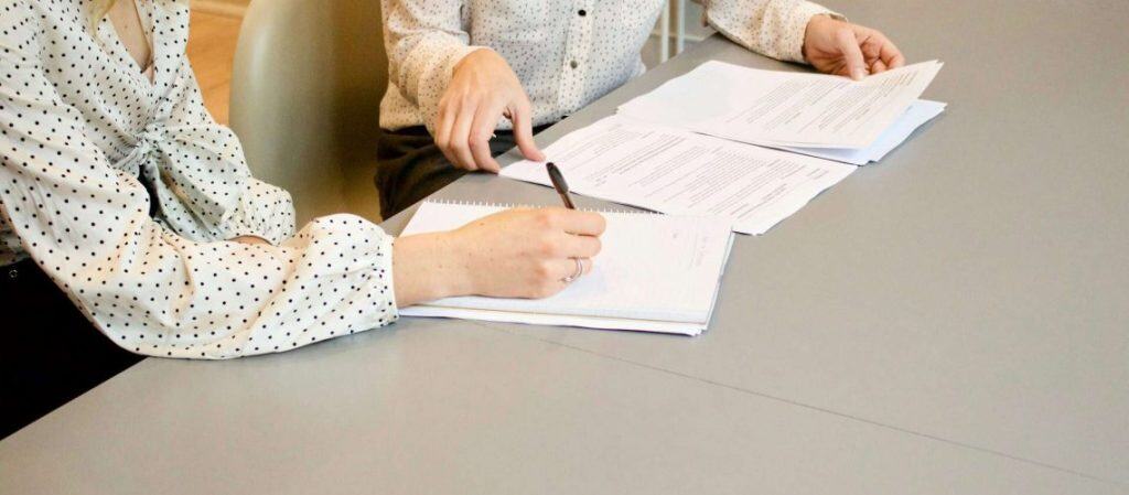 Two people wearing polka dot shirts are sitting at a table reviewing documents. One points to a page, while the other holds a pen, indicating collaboration.