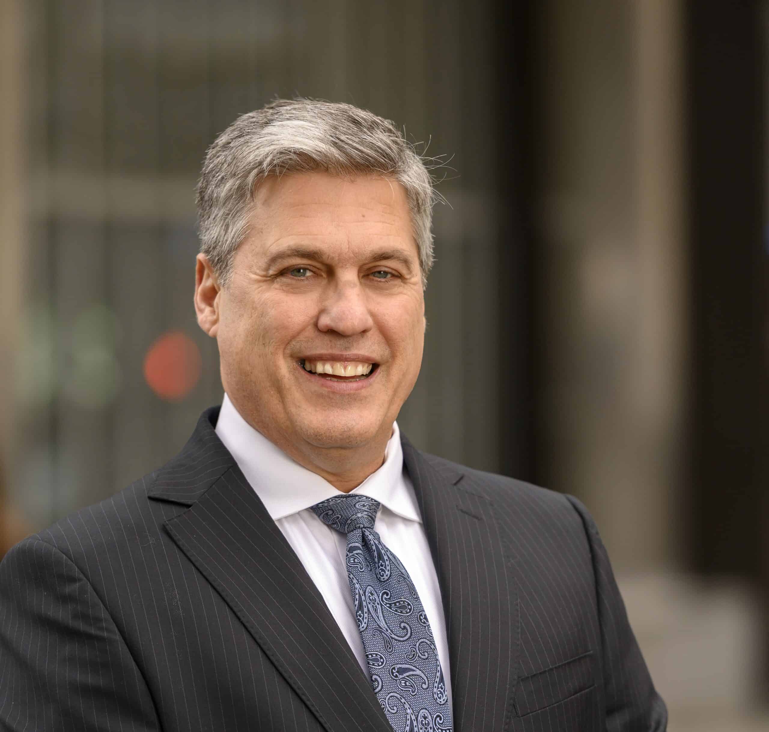 A smiling man in a suit and patterned tie stands outdoors. The background is a blurred urban setting.