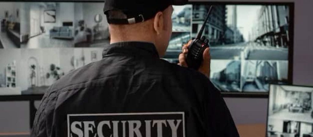 Security personnel in uniform monitoring multiple surveillance screens, holding a walkie-talkie, in a control room.