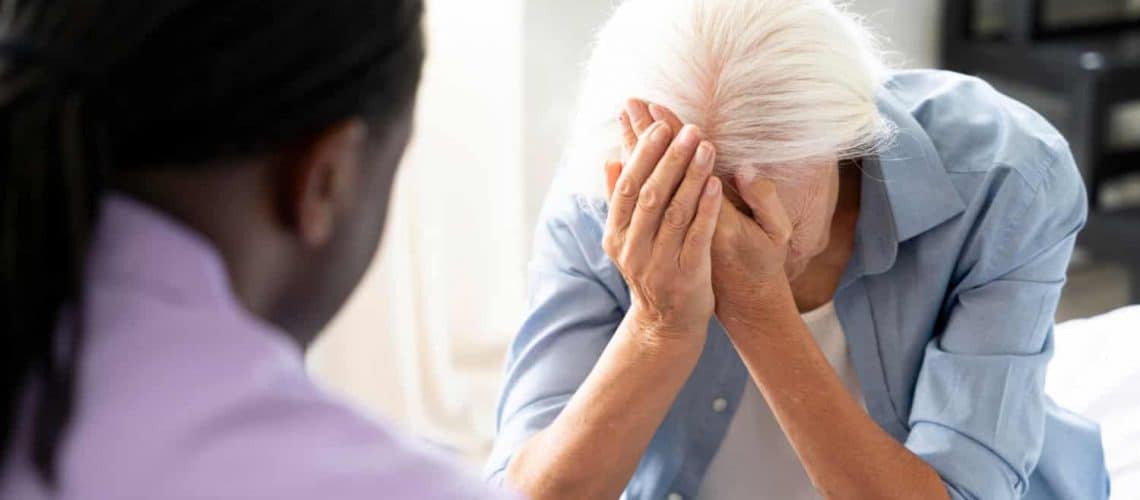 An elderly person sitting with their head in hands, appearing distressed, while another person sits nearby, offering support.