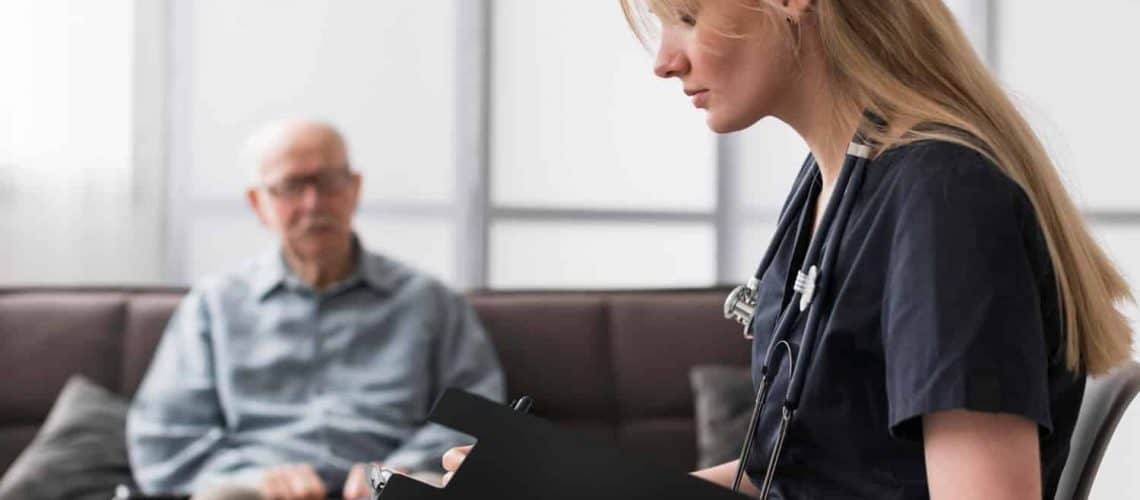 A healthcare professional with a stethoscope and clipboard talks to an elderly man sitting on a sofa in an office setting.