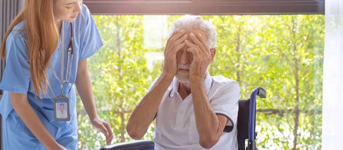 A nurse tends to a man in a wheelchair, who is covering his face with his hands. Bright window with greenery in the background.
