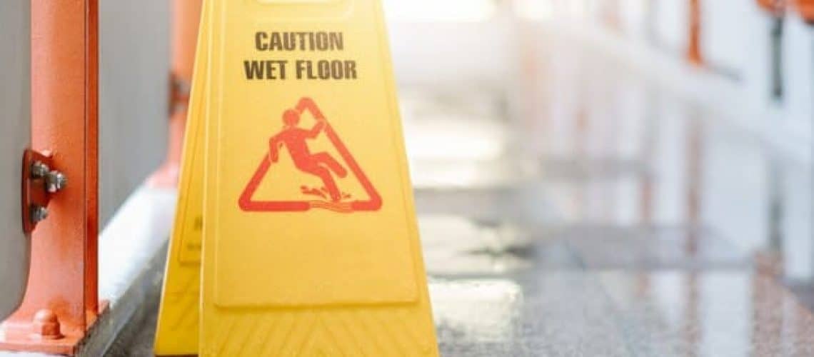 Yellow "Caution Wet Floor" sign on a shiny tiled floor with sunlight in the background.