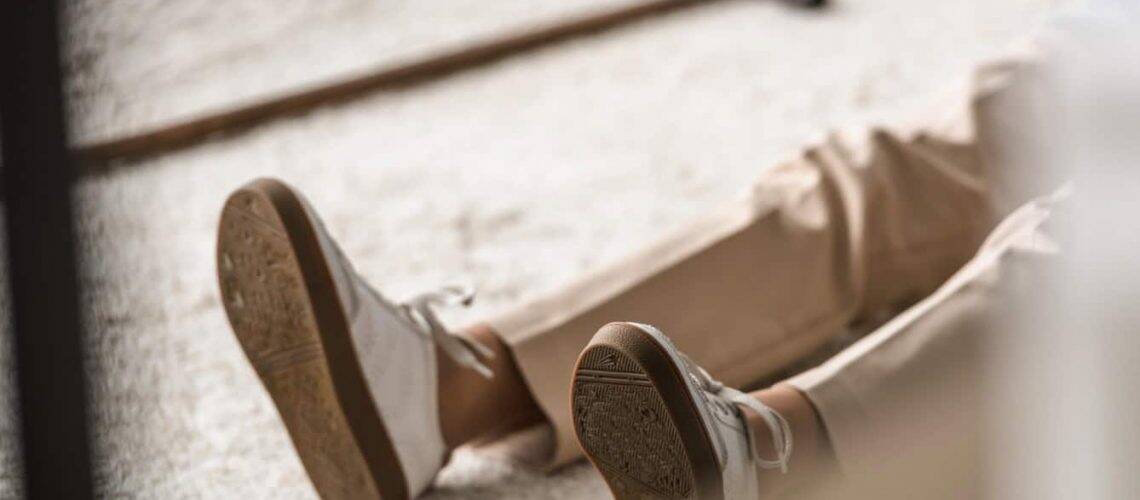 Person lying on a carpeted floor with legs visible, wearing light-colored pants and white sneakers.