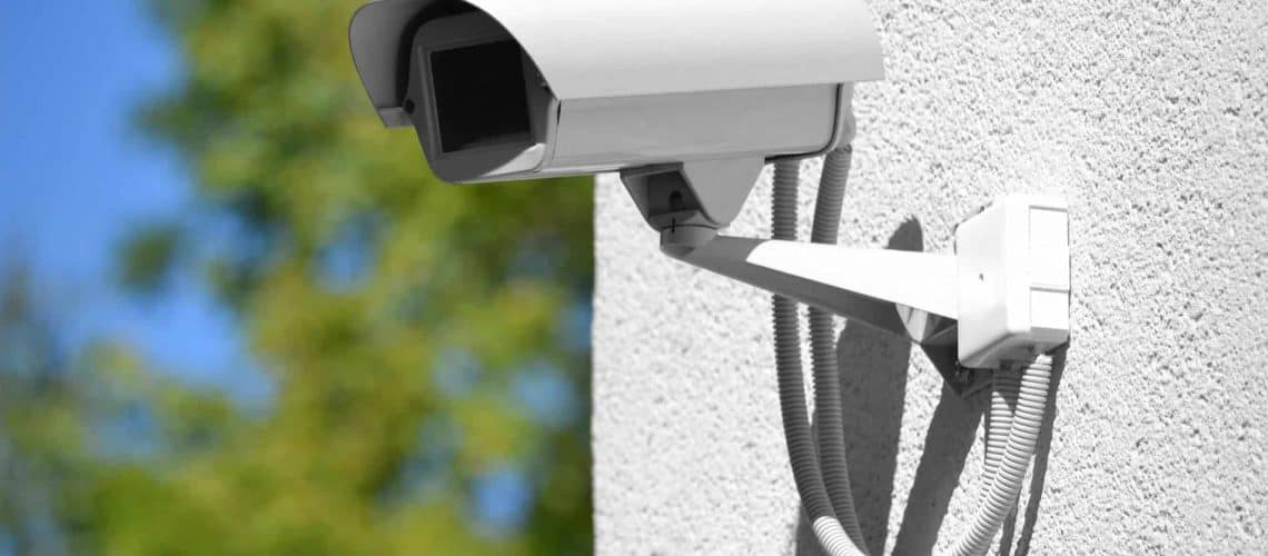 A white security camera mounted on a textured wall with cables visible, set against a blurred green background.