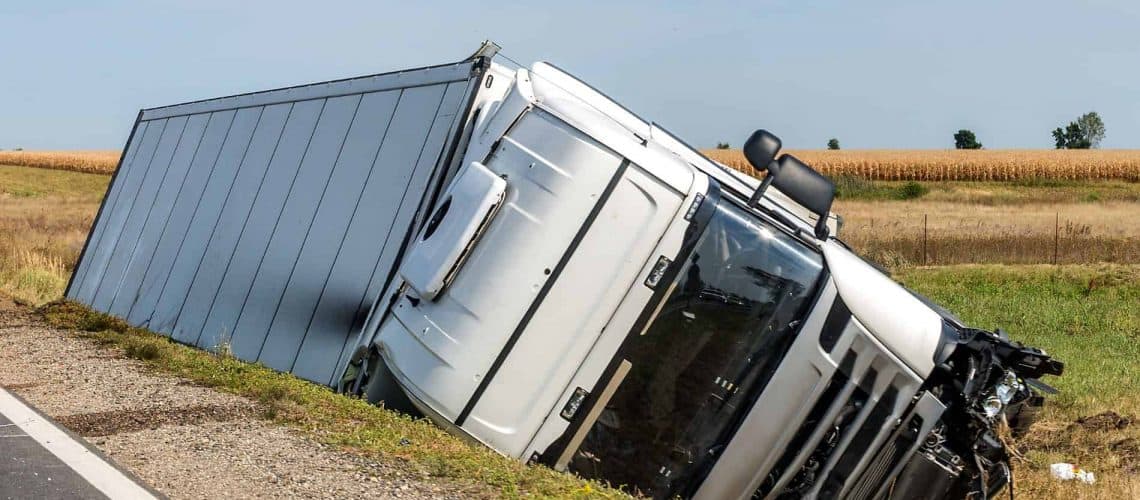 A white trailer truck is tipped over into a roadside ditch on a rural highway, with fields and clear sky in the background.