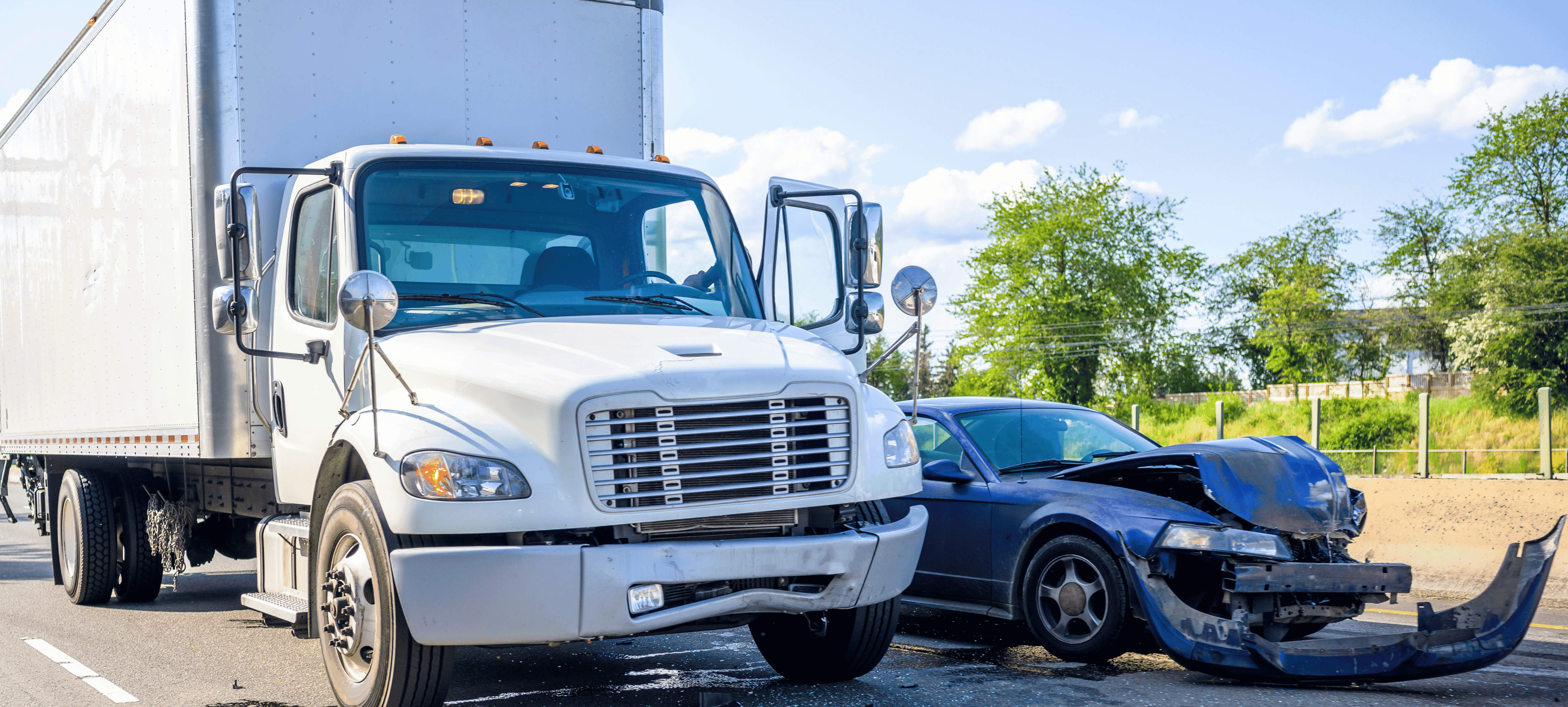 White semi-truck and blue car in a collision on a road. Car's front is damaged. Trees and fence in the background.