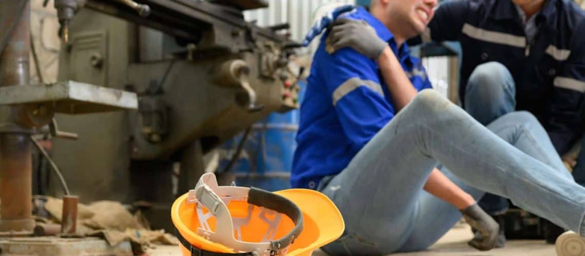 A person in a blue shirt sitting on the floor of an industrial space appears injured, holding their shoulder. Another person kneels beside them. A yellow hard hat lies on the ground nearby.