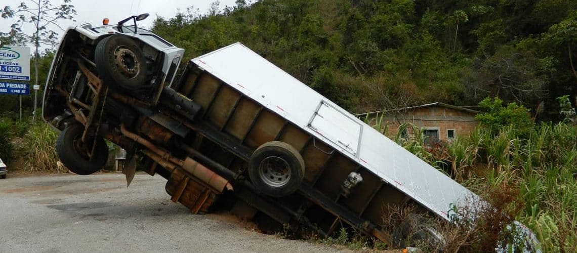 A truck is tipped upward into the air at an angle, with its front wheels off the ground, on a roadside near vegetation and a small building.