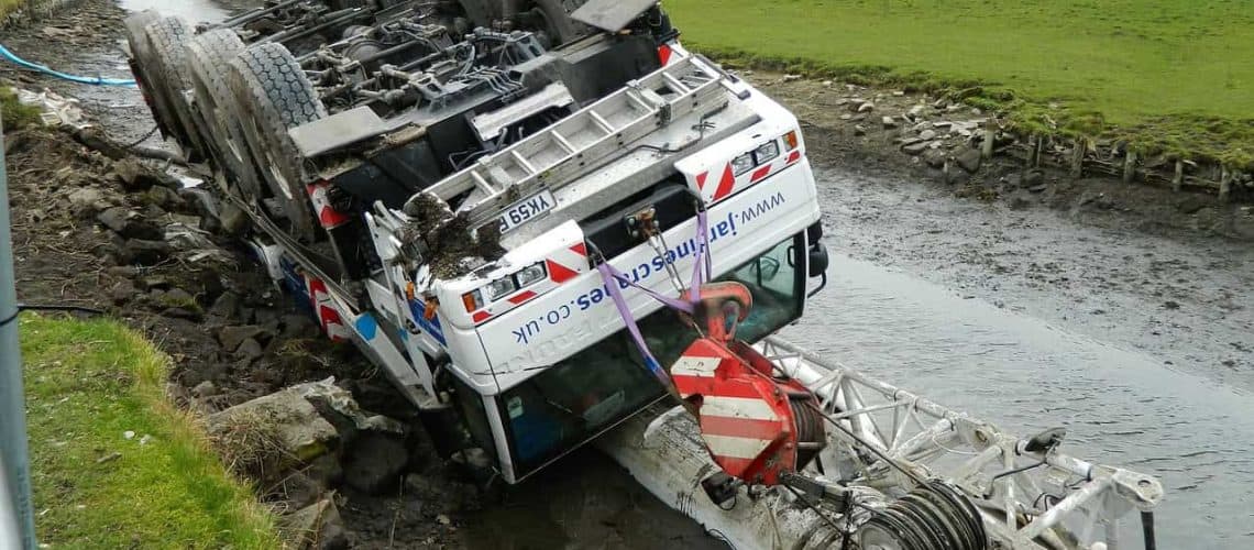 A large vehicle overturned on its side in a rocky, muddy area near a body of water, with machinery partially submerged.