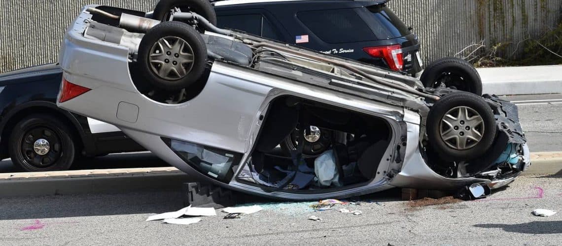 Overturned silver car on a city street after an accident, with debris scattered around. A black SUV is parked nearby.