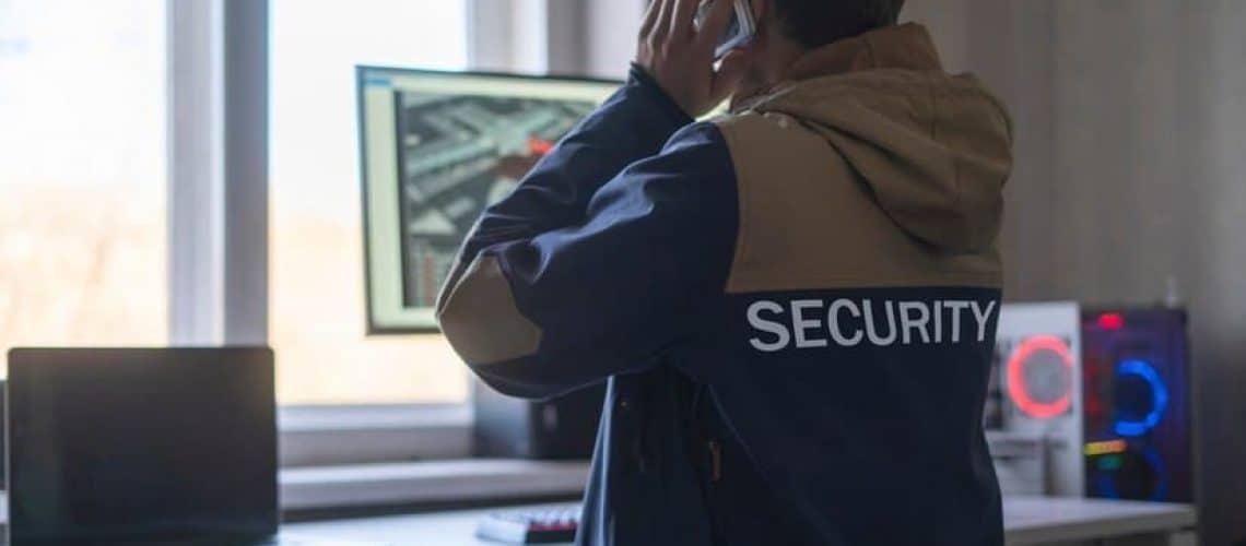 Person wearing a security jacket is using a smartphone while looking at computer screens in a room with windows.