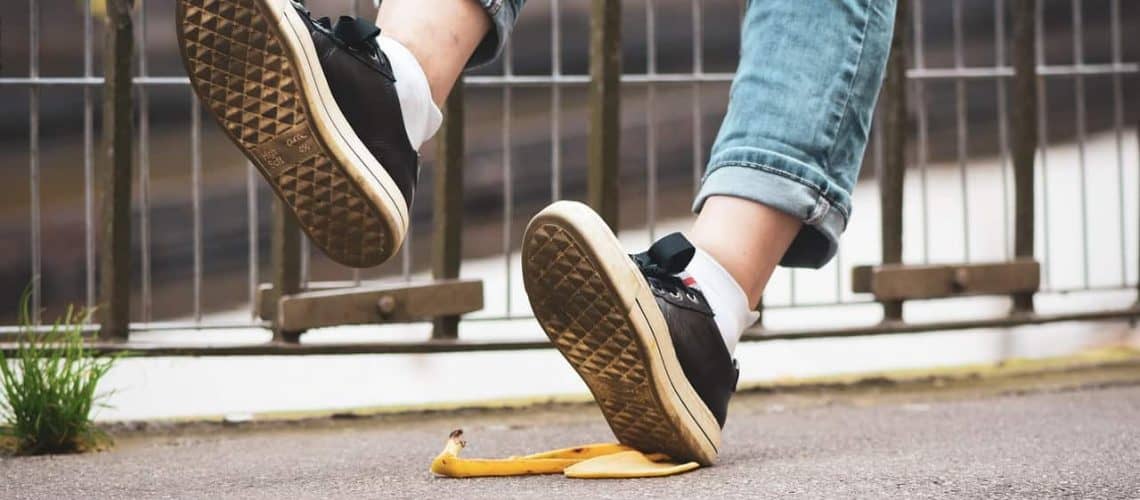 Person slipping on a banana peel on the pavement, wearing rolled-up jeans and black sneakers.