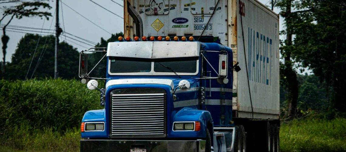 A blue semi-truck with white trailer drives on a rural road, surrounded by trees and grass.