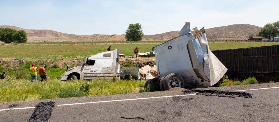 Overturned white semi-truck off the road in a grassy area. People in high-visibility vests assess the scene under a clear sky.
