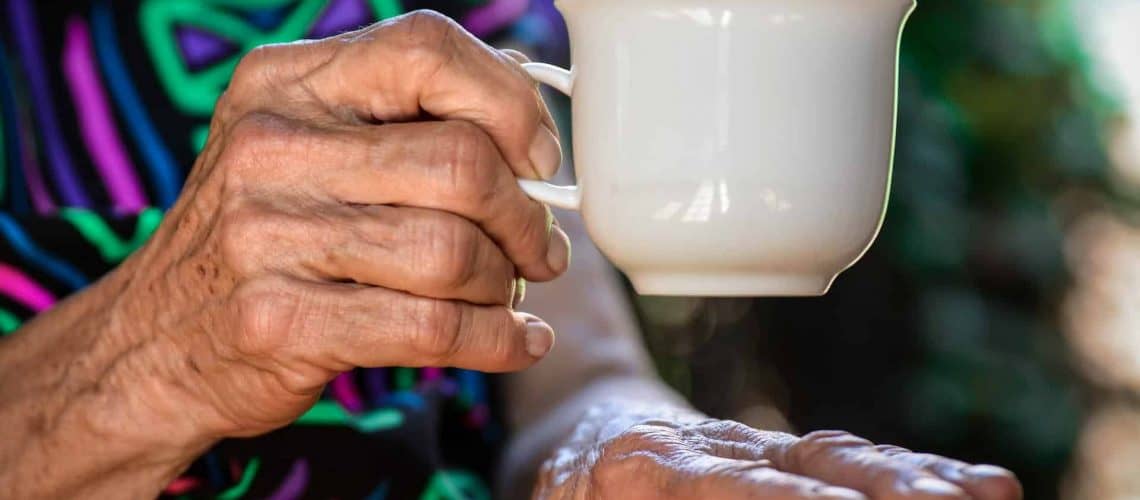 Elderly hands holding a white cup, with a colorful patterned garment in the background.