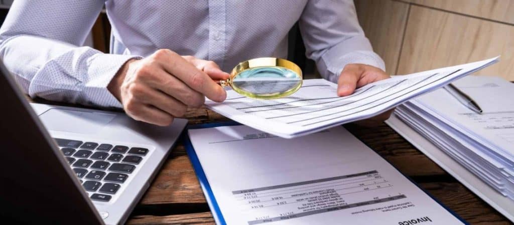 Person examining a document with a magnifying glass at a desk, alongside a laptop, invoice, and clipboard.