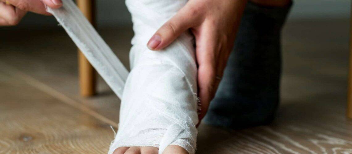 A person wrapping a bandage around their foot and ankle while seated on a wooden floor.