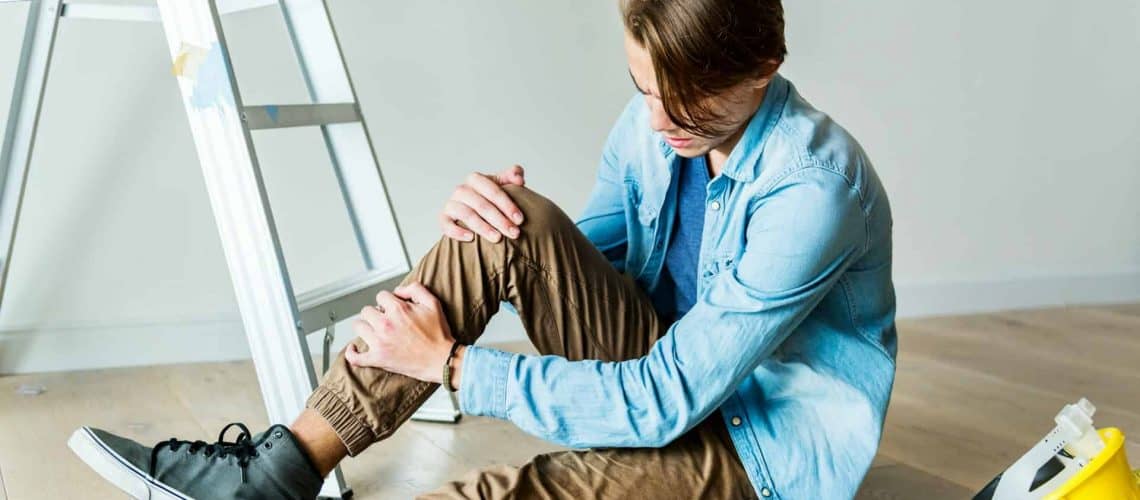 A person sitting on the floor holding their knee, with an overturned ladder and a toolbox nearby.