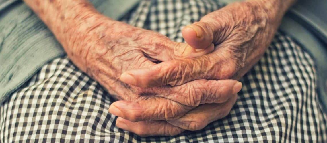Elderly hands with crossed fingers rest on a black and white checkered fabric.