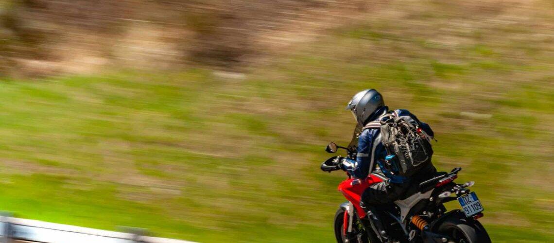 Motorcyclist wearing a helmet and backpack rides a red and black motorcycle on a road, with blurred green landscape in the background.