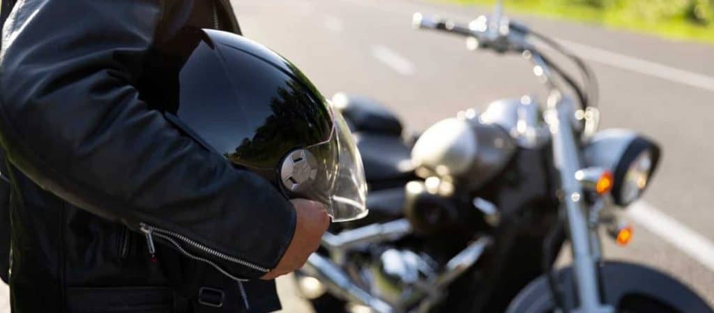 A person in a leather jacket holds a black motorcycle helmet near a parked motorcycle on an asphalt road.