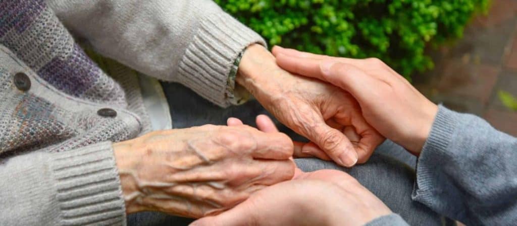 An elderly person wearing a sweater is holding hands with a younger person. Greenery is visible in the background.