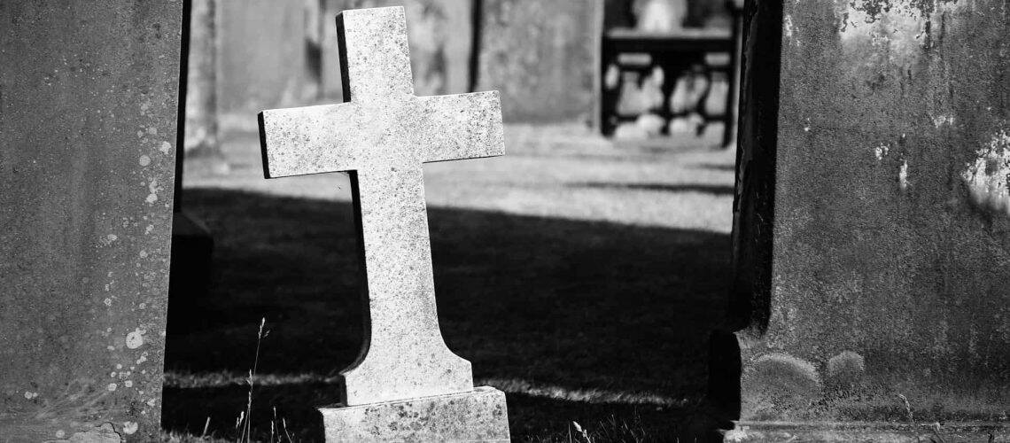 A leaning stone cross in a graveyard, surrounded by other grave markers, in black and white.
