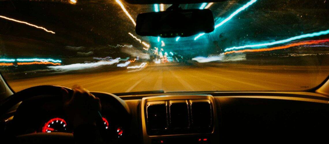 View from inside a moving car at night, showing a blurry street with lights streaking past, suggesting speed. Dashboard and steering wheel visible.