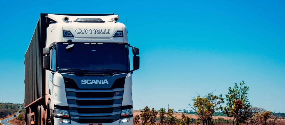 A white Scania truck with "Comelli" on the front drives on a rural road, with blue sky and trees in the background.