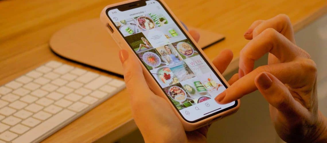 Person scrolling on a smartphone, viewing a social media profile featuring various food and lifestyle images, with a keyboard in the background.
