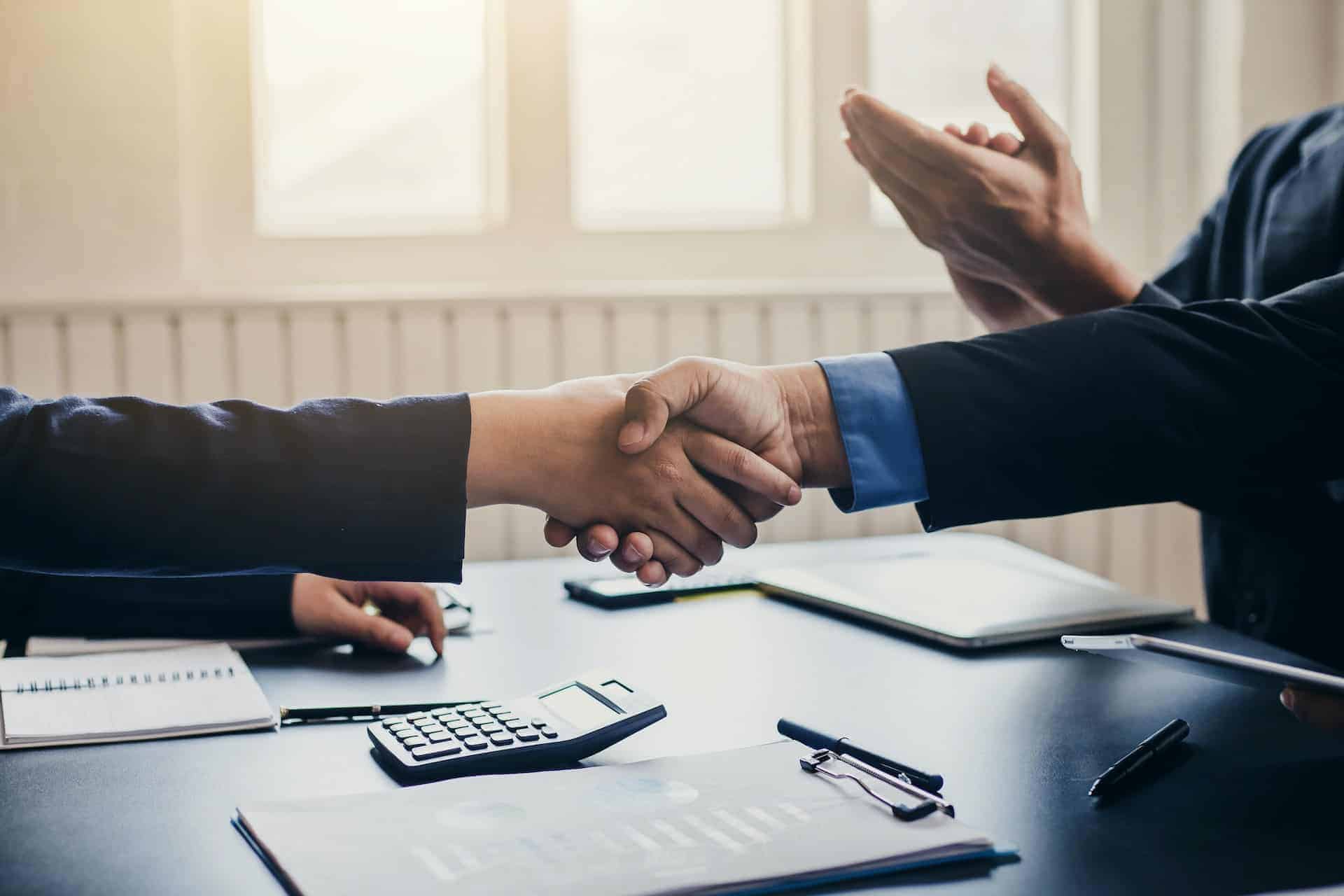 Two people in business attire shaking hands over a table. A third person is clapping. A calculator, documents, and laptops are visible on the table.