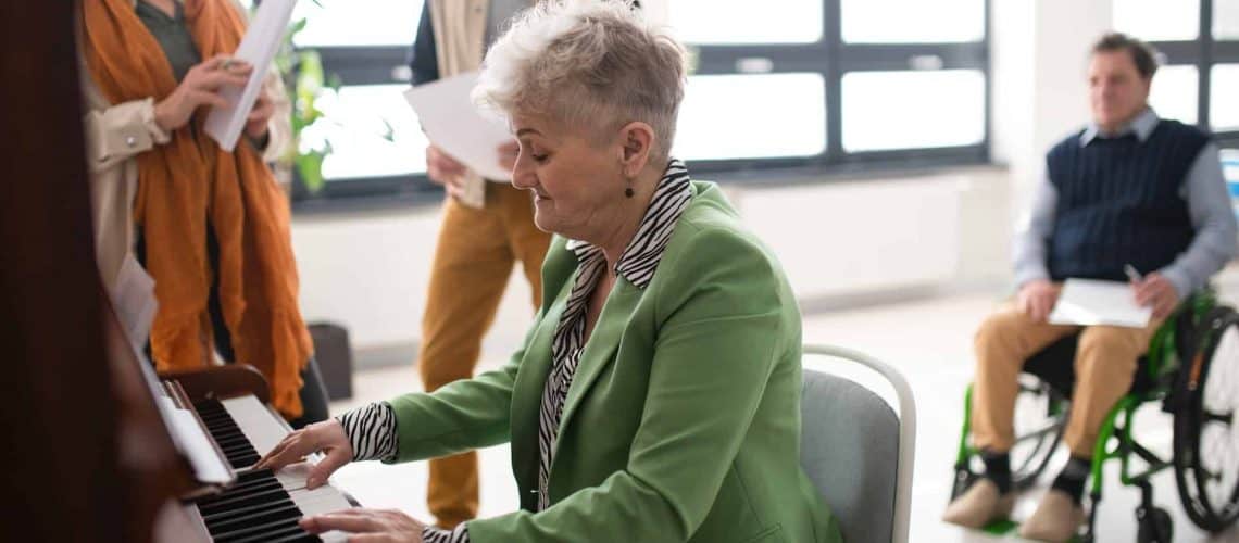 Older woman in green blazer playing piano in a bright room. A group of people stands nearby and one person in a wheelchair observes.