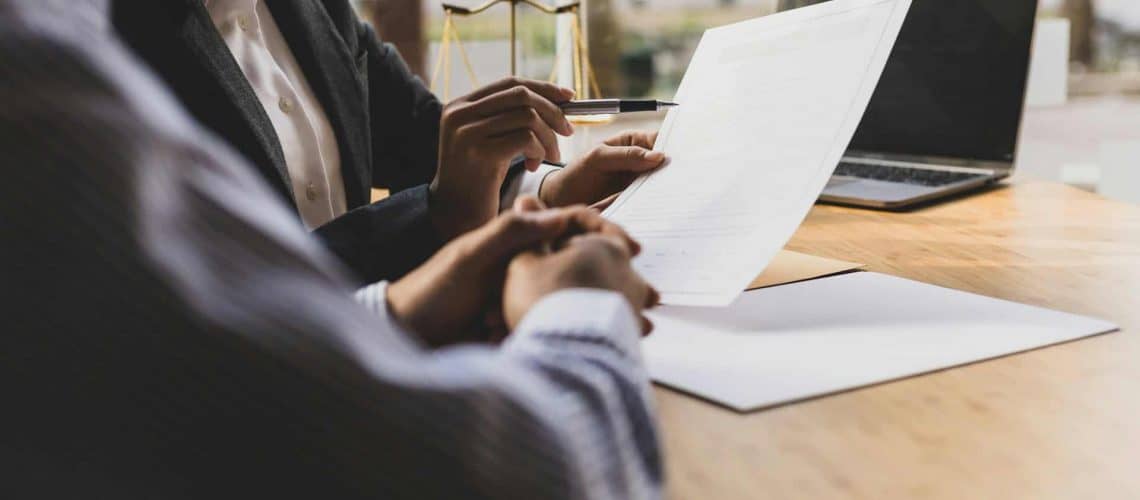 Two people in business attire review a document at a table with a laptop and notepad nearby.